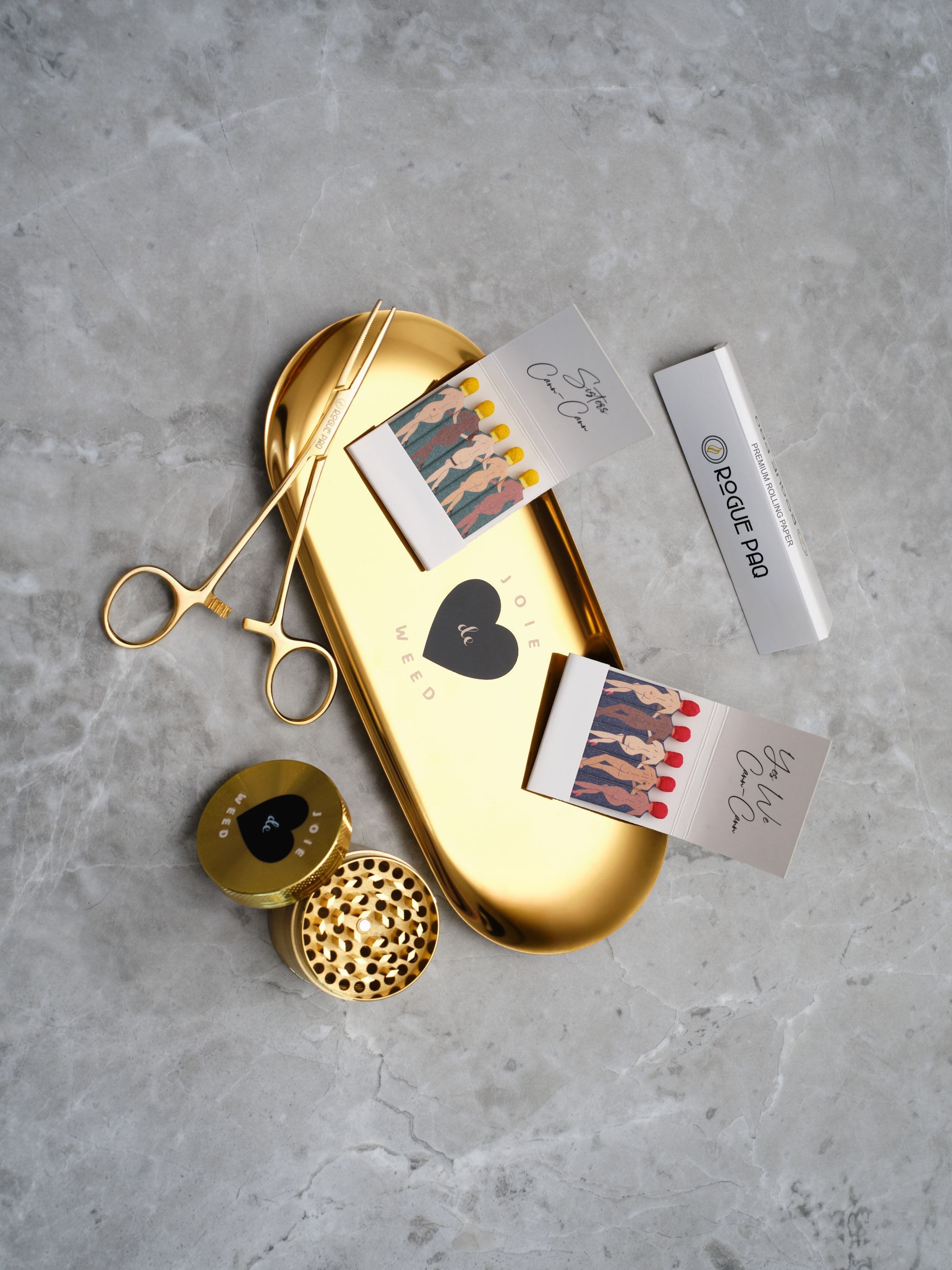 A luxury cannabis gift set featuring a booklet of Rogue Paq unbleached hemp rolling papers, Eiffel Tower Scissors, a refillable lighter, two books of SendNudes matches, and a pair of gold-toned hemostat jay clips. The set is presented in a minimalist, high-contrast arrangement that highlights the polished gold hardware.