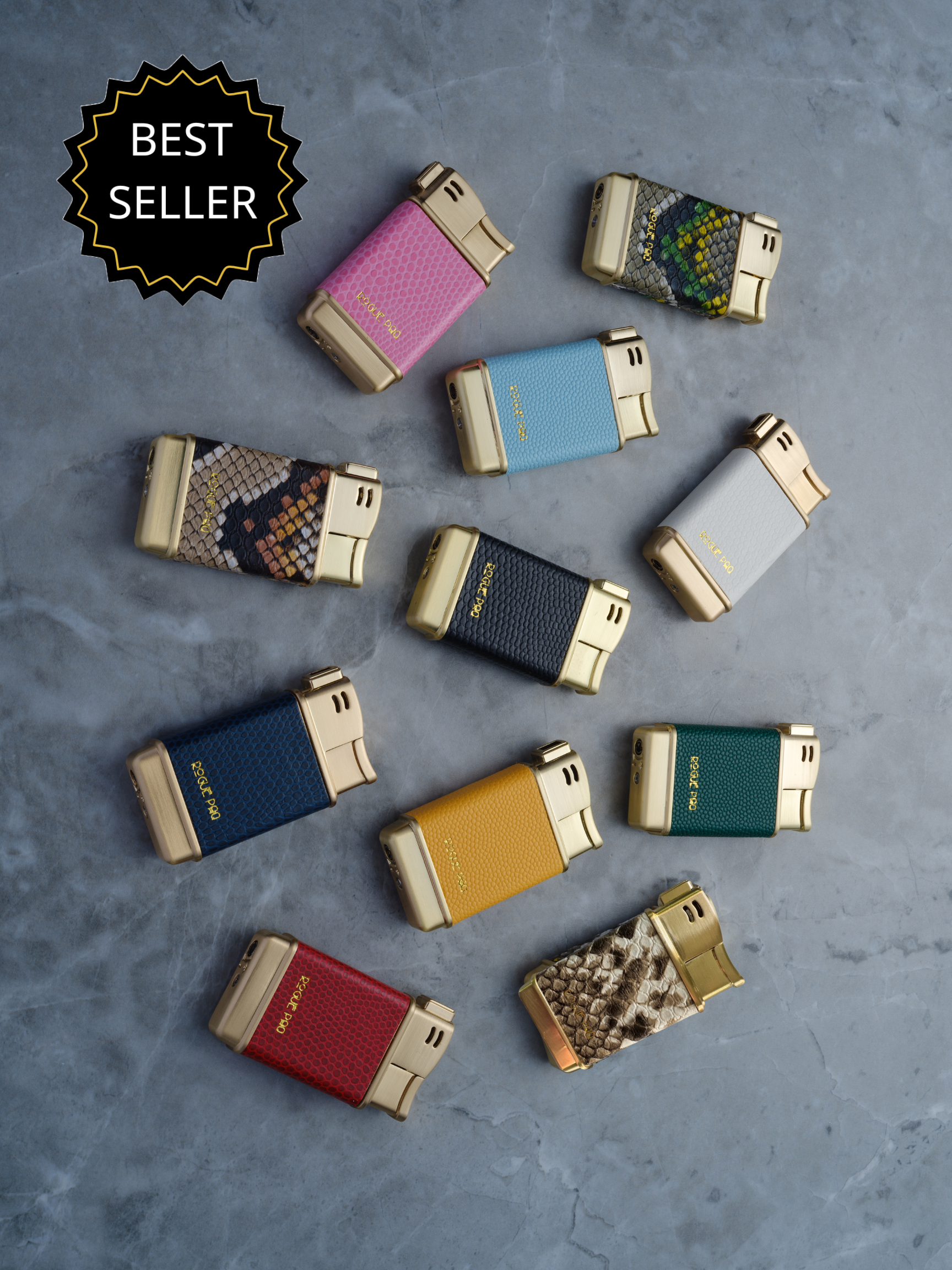 A vibrant spread of Rogue Paq refillable butane lighters in a variety of colors and textures, including vegan lizard skin and vegan snakeskin. The lighters are arranged to showcase the high-shine gold-tone casing and the 'Raise Your Ritual' aesthetic. They are the epitome of luxury smoking accessories.