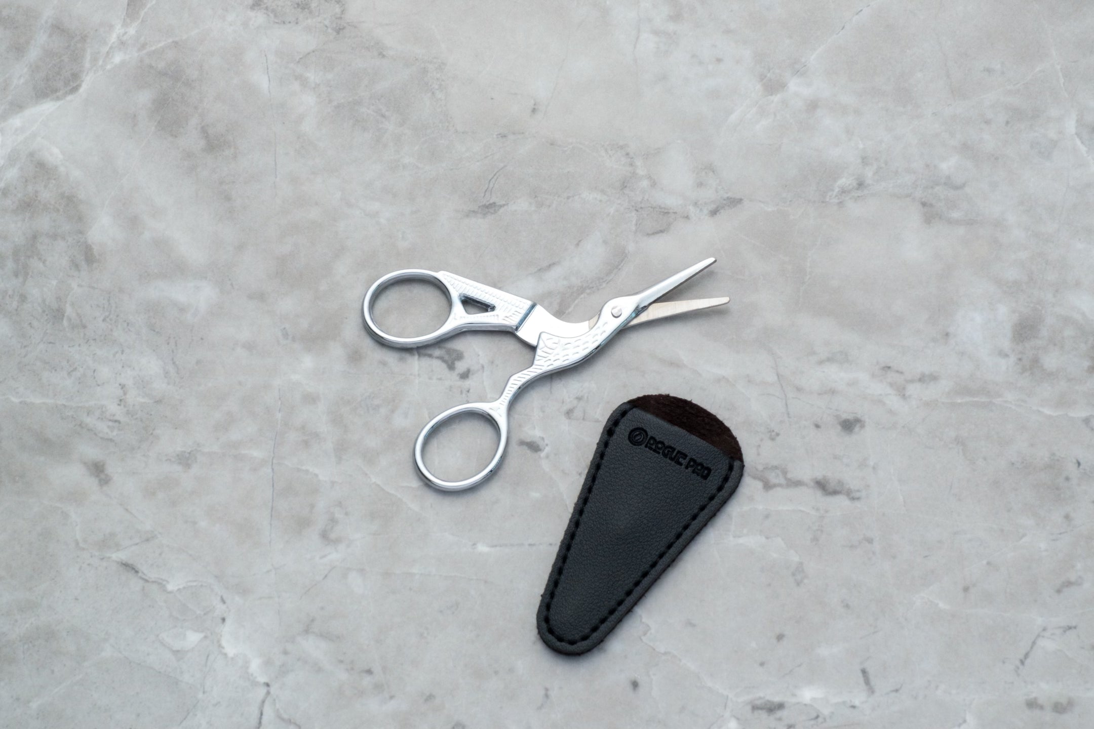 A pair of silver-toned precision trim scissors designed in the shape of a crane. The body of the bird forms the handles, while the long, slender beak serves as the sharp, stainless steel blades, finished in a high-polish silver for a luxury aesthetic. They are pictured on a gray marble background along with their black vegan leather blade cover.