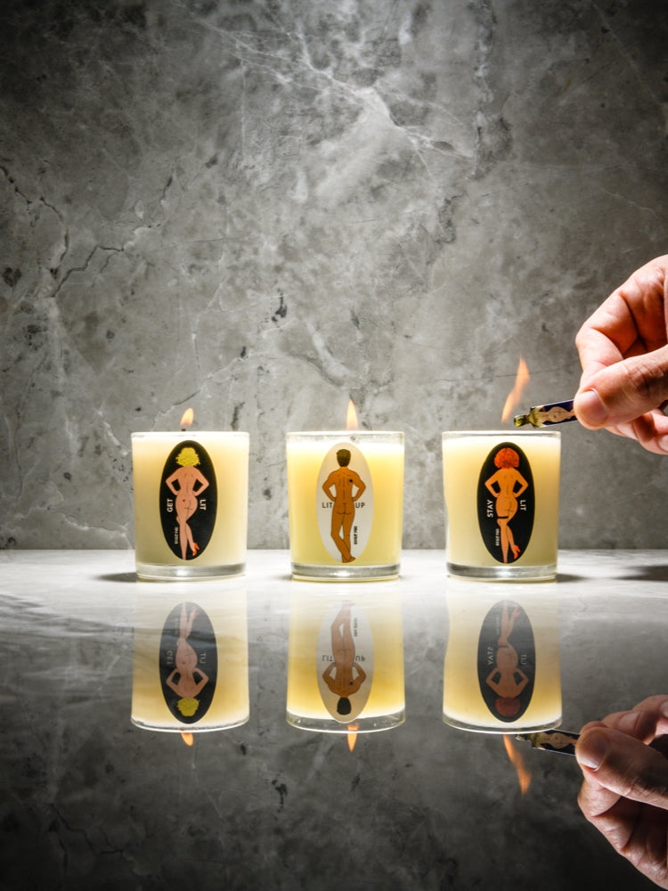 A curated set of three 2oz Rogue Paq organic massage candles—Get Lit, Stay Lit, and Lit Up. The clear glass votives are arranged in a clean, sophisticated row against a marble background. They are being lit with a SendNudes match.