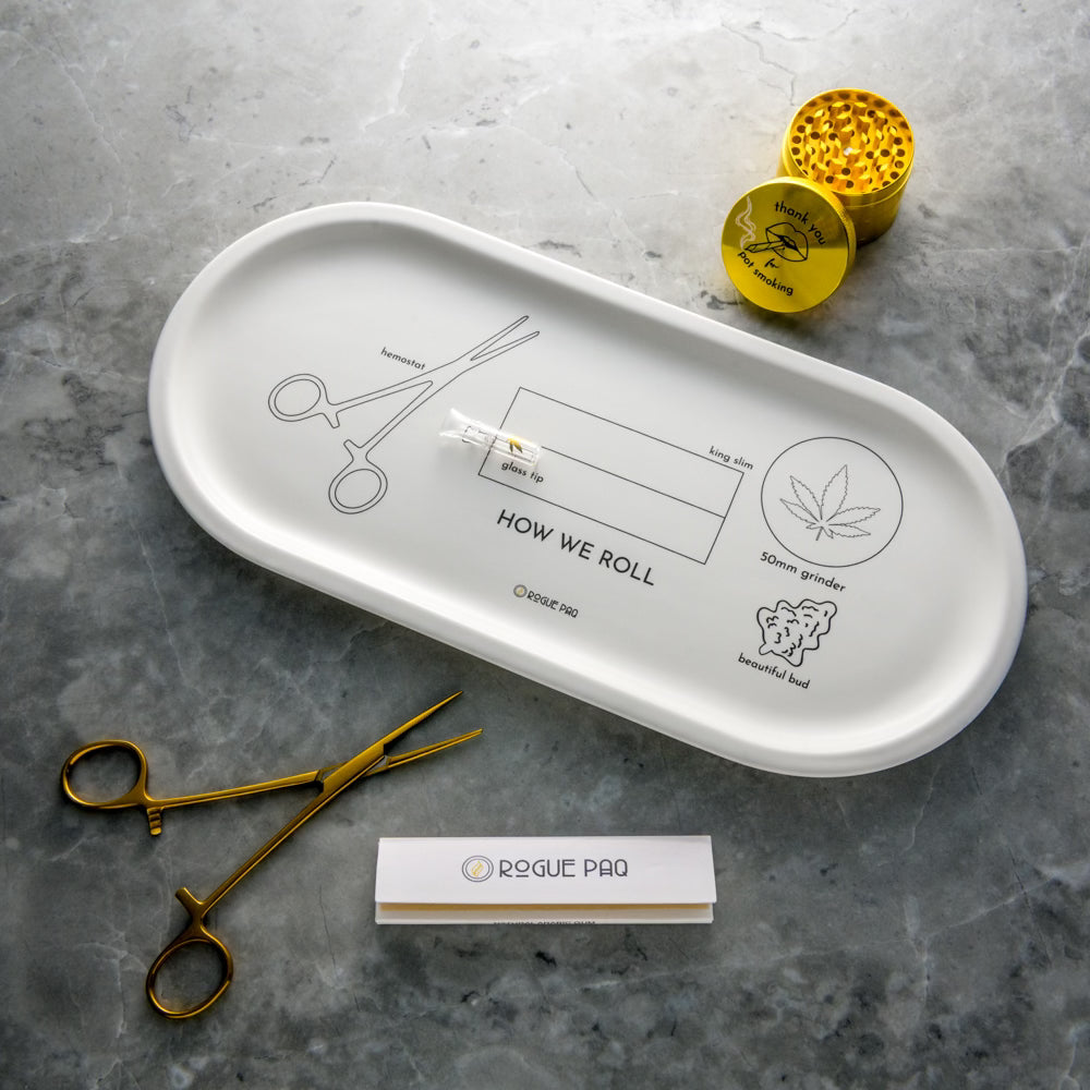 Rogue Paq How We Roll Gift Set featuring the white porcelain valet tray with instructional rolling illustrations; styled with a gold-tone grinder, hemostat clip, glass tip, and hemp rolling papers.