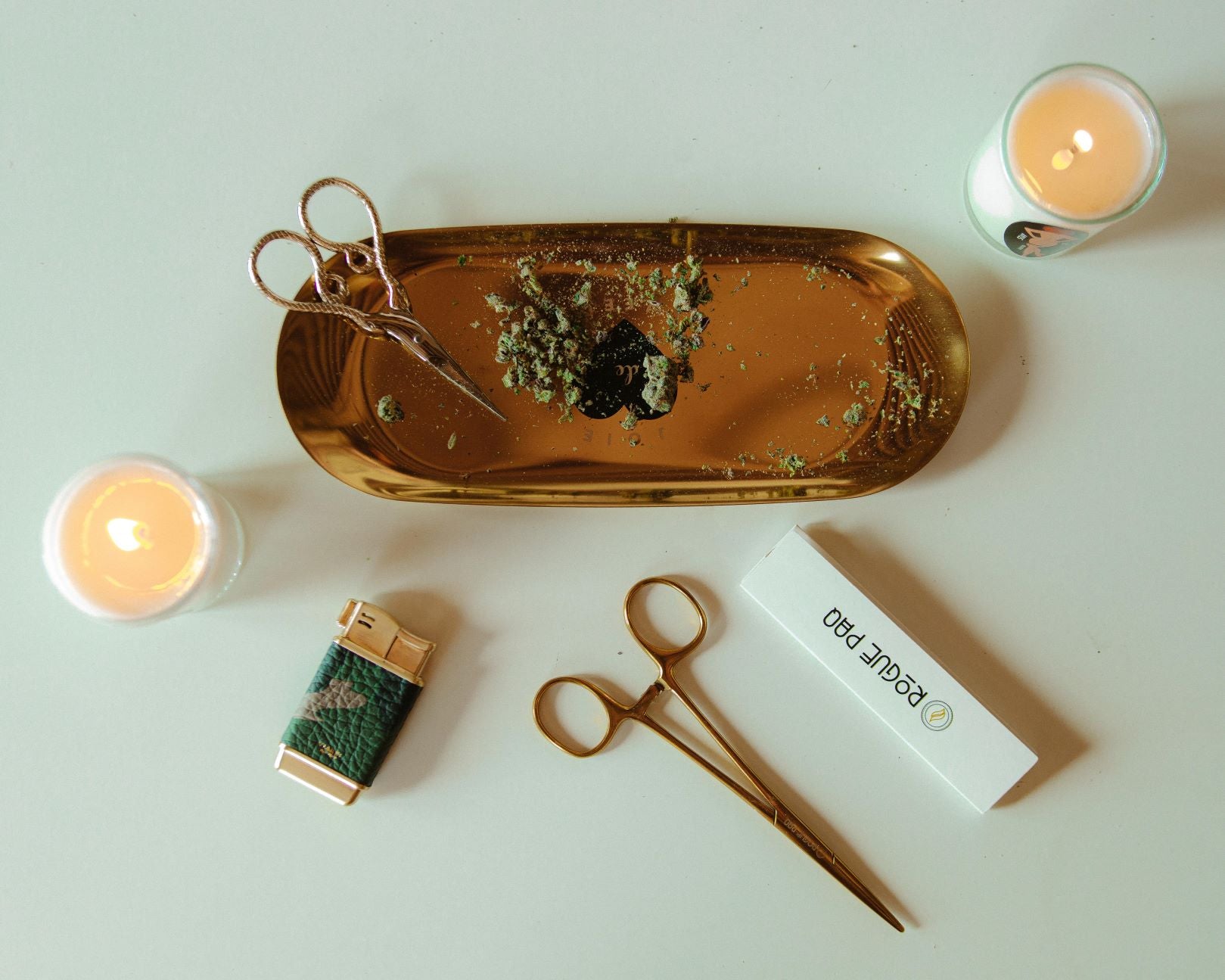 A lifestyle image of the gold python scissors on the Rogue Paq Joie de Weed travel rolling tray, demonstrating their compact and ergonomic scale. The light catches the textured gold handles, illustrating how the tool functions as both a high-utility instrument and a statement accessory. Also pictured is a SendNudes candle, book of rolling papers, a hemostat joint clip and a refillable lighter.