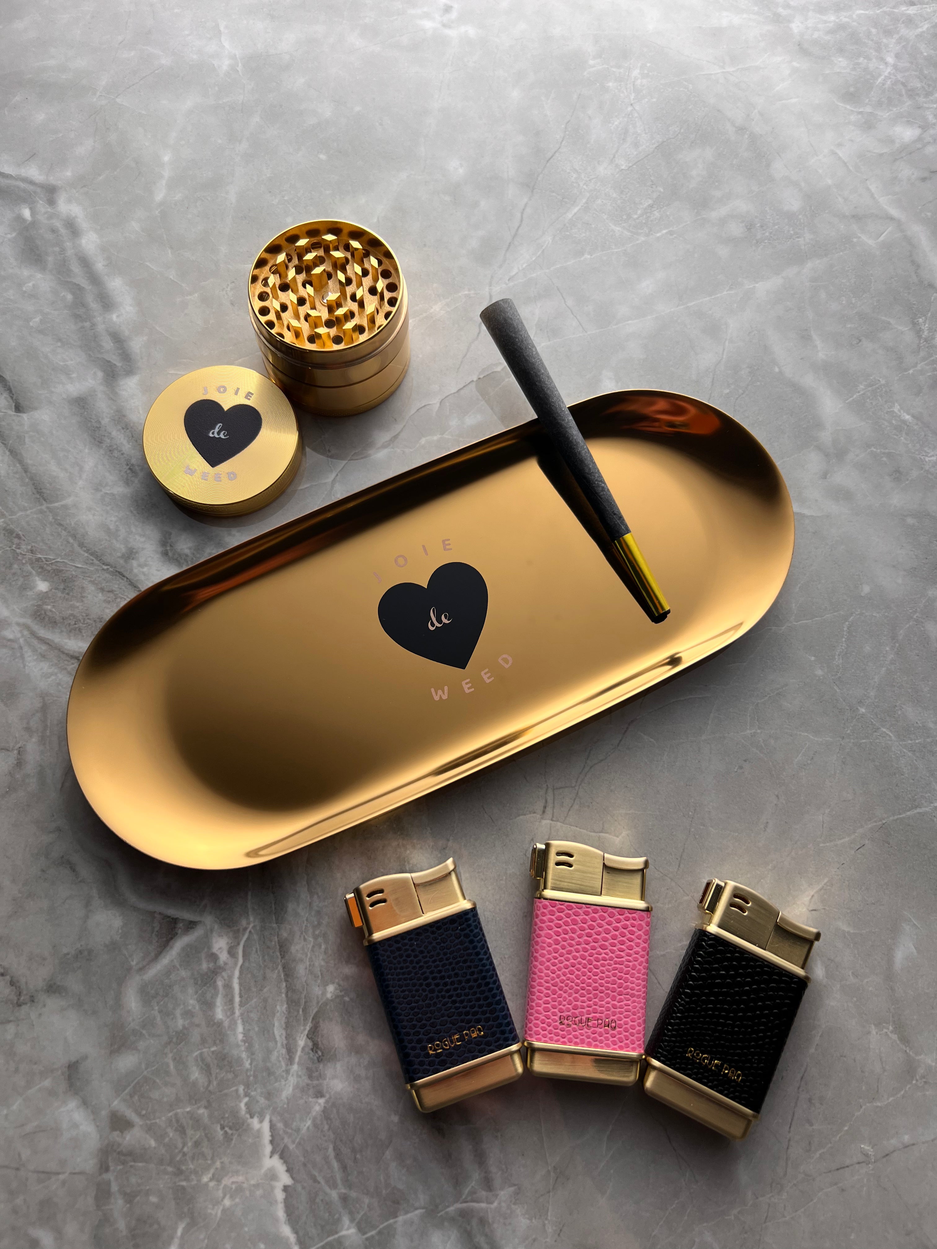 A lifestyle shot of the Rogue Paq Joie de Weed travel rolling tray and grinder. The tray holds a Rogue Paq all natural hemp cone. The shot includes three refillable butane lighters in caviar, bubble gum, and marine. All of the products are set on a gray marble surface.