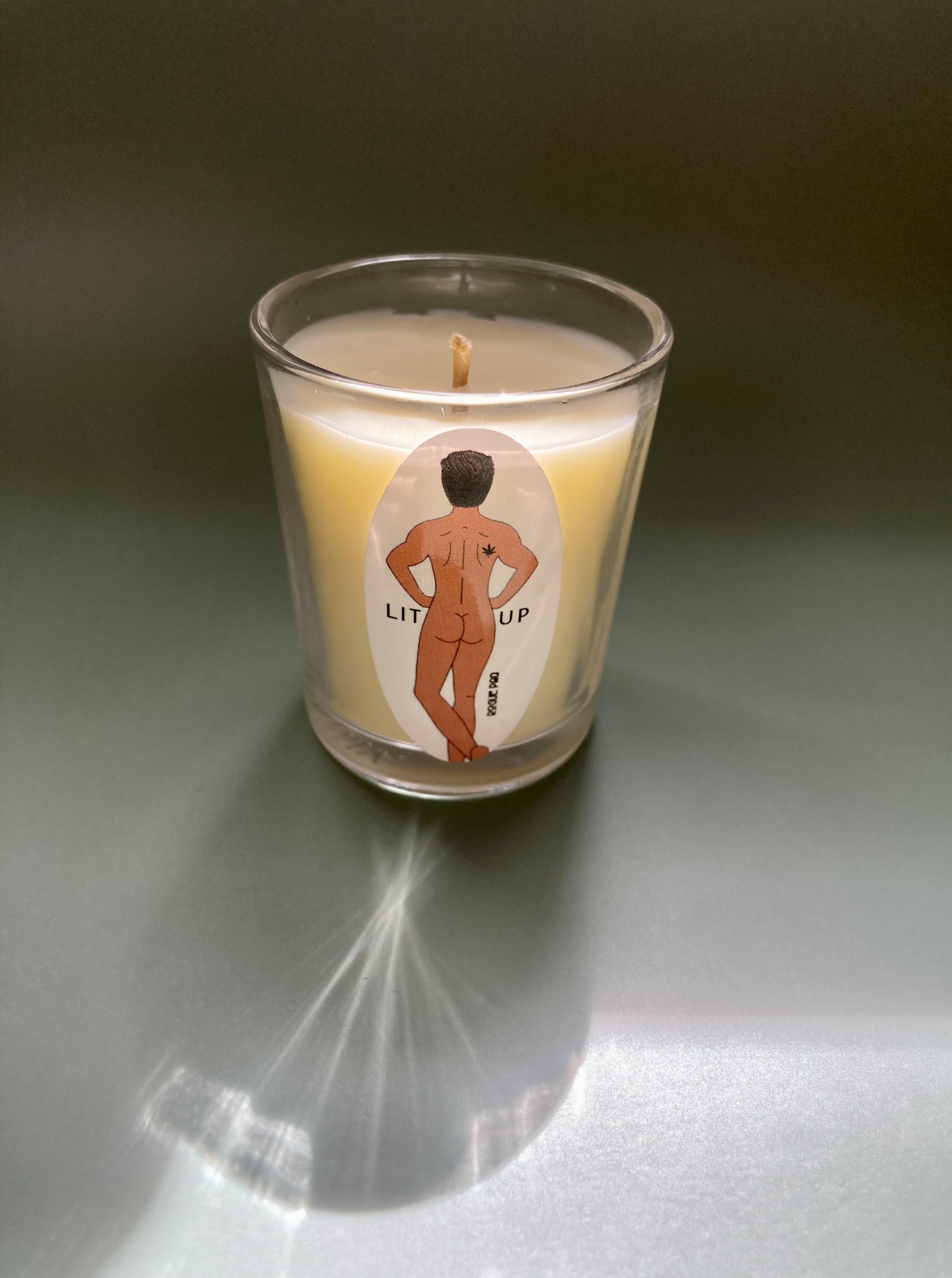 A close-up shot of the 2oz LIT UP organic soy massage candle. The natural, off-white wax is hand-poured into a clear glass votive with a rustic hemp wick. The clean label emphasizes the uplifting aromatherapy blend, designed for both scent and skin-safe massage oil use. The label features the signature Rogue Paq can-can nude dancer which is a male figure with black hair and light brown skin.
