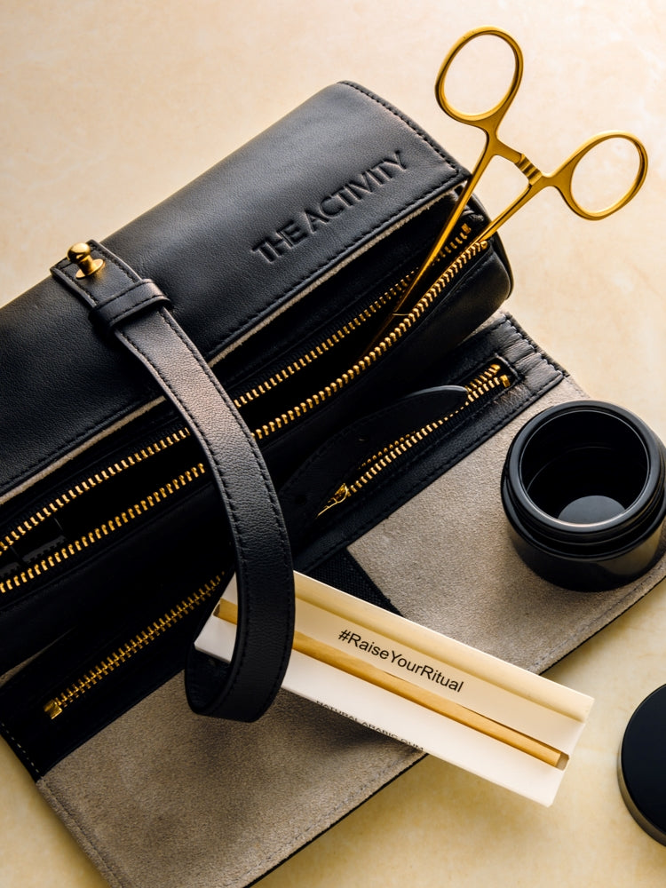 A black lambskin leather cannabis carrying case with gold-toned hardware, displayed alongside a jar, hemostats and other accessories. The signature Rogue Paq Ritual Case is shown here in premium black lambskin leather. The case is shown in its signature rolled position, with its wrap-around leather strap undone. The gold-toned hardware is highlighted presenting as a sleek, discreet, and luxury travel accessory.