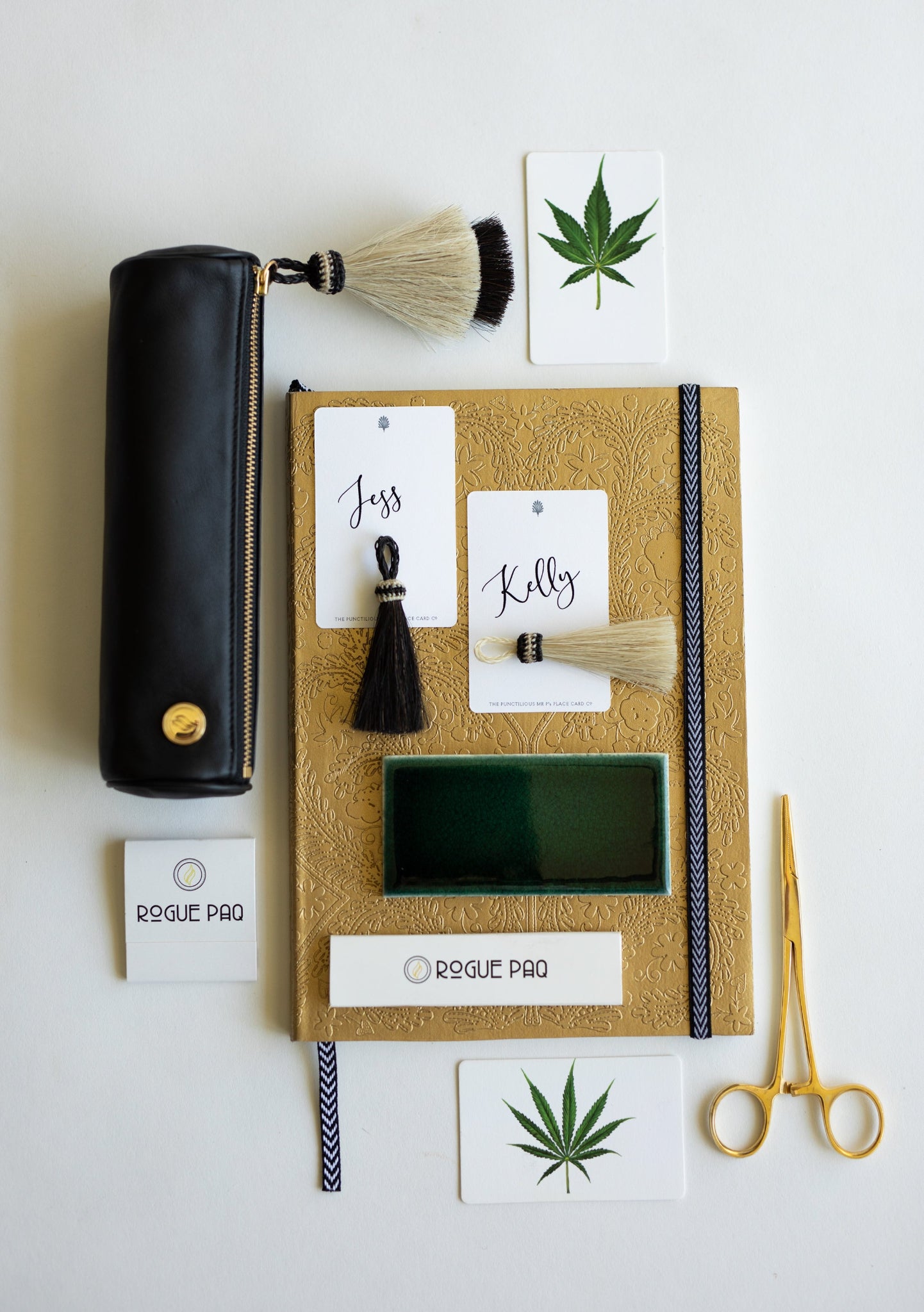 A professional lifestyle flatlay on a white office desk featuring the Rogue Paq x KellyGreen cannabis case. The black leather case is paired with a matching horsehair tassel, stationery, and a gold-toned hemostat, illustrating its seamless integration into a polished, modern aesthetic.