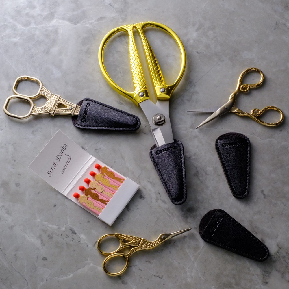 4 styles of Rogue Paq scissors on a gray marble background. All Scissors come with a vegan leather blade cover for easy transport and safety. The shot includes a book of Rogue Paq best selling SendNudes vintage inspired matches with nude dancing girls and the text Send Doobs.