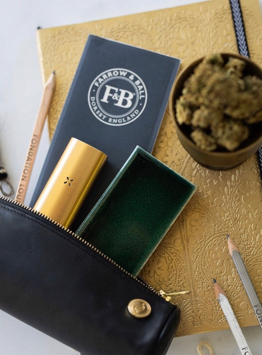 The Solo Paq ritual case shown alongside a cup of premium cannabis bud and a gold PAX. This close-up highlights the fine-grained leather and the gold zipper detail, illustrating a high-end approach to the smoking ritual.