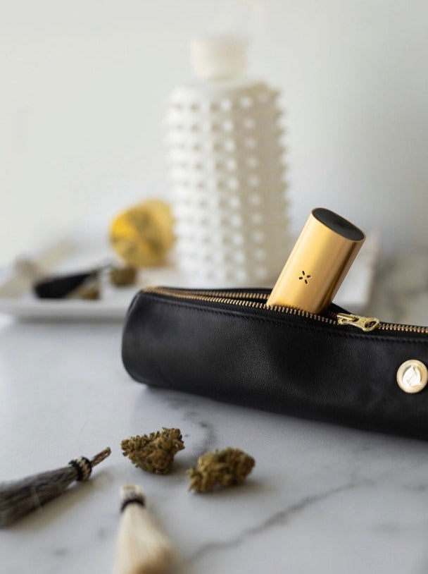 A lifestyle shot of the black leather Solo Paq case paired with a PAX vaporizer. The image emphasizes the buttery texture of the lambskin and the luxury appeal of the brand's coordinated cannabis accessories.