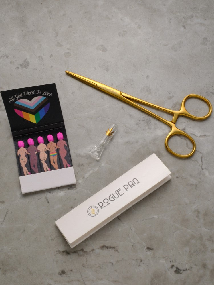 A lead shot of the Rogue Paq Bootyful (Pride) Bundle. The set includes a pair of gold-toned hemostat jay clips, a booklet of Rogue Paq unbleached hemp rolling papers, a glass filter tip with a gold cannabis leaf decal, and an 'All You Weed Is Love' matchbook featuring inclusive dancing figures.