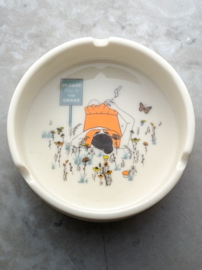 Round porcelain ashtray featuring the whimsical phrase 'Please Keep On The Grass' in white lettering; a playful and elegant home accessory for the cannabis enthusiast