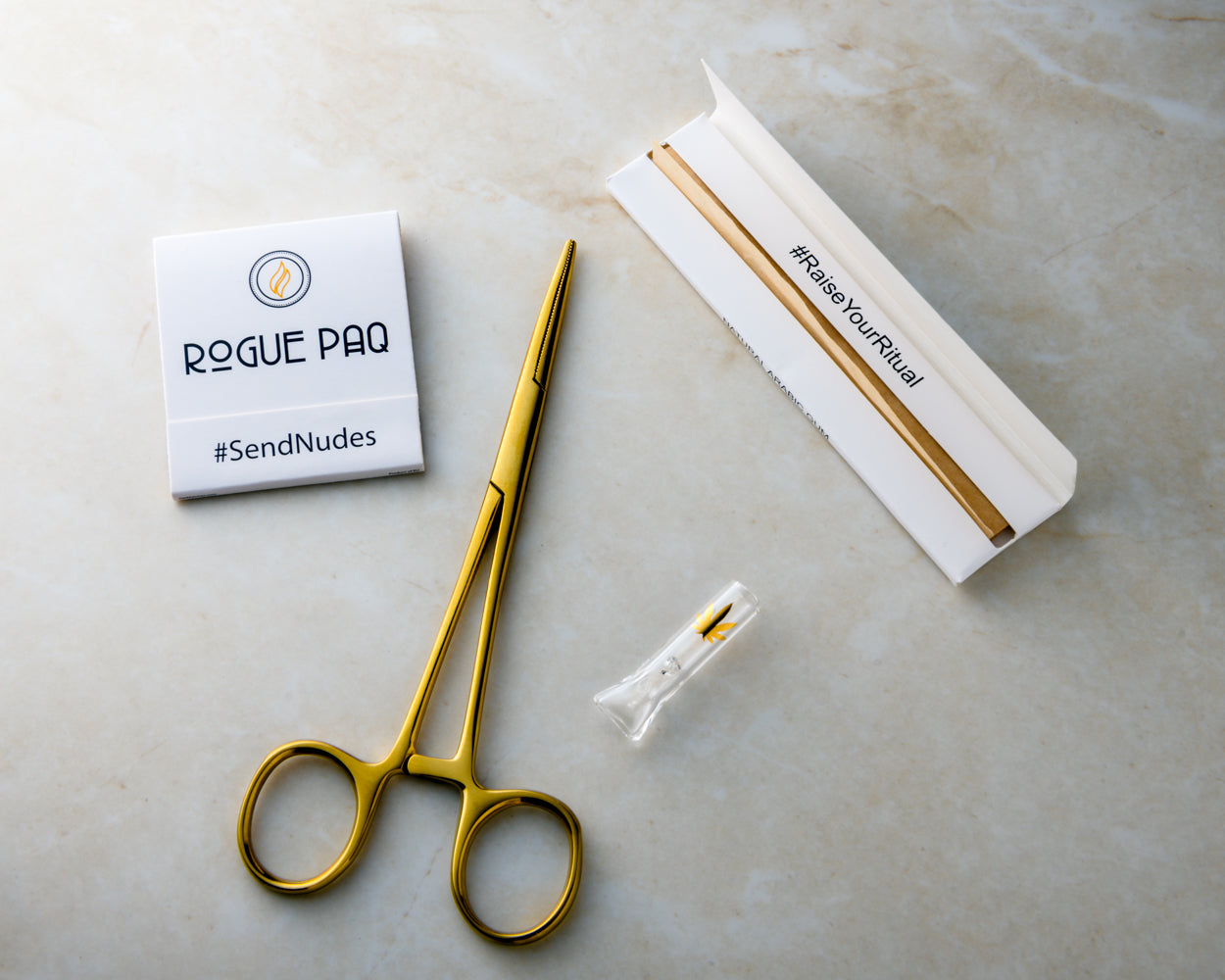 A detailed group shot of the Black Lives Matter Give Back Bundle. The set features gold-toned hemostat packing tools, unbleached hemp rolling papers, a glass tip with a gold-leaf accent, and a signature 'SendNudes' matchbook. 100% of proceeds from this bundle are donated to social justice organizations.