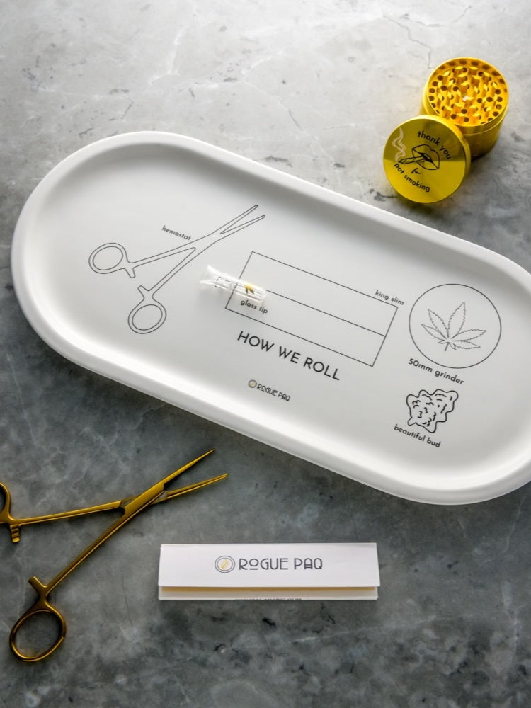 Rogue Paq How We Roll Gift Set featuring the white porcelain valet tray with instructional rolling illustrations; styled with a gold-tone grinder, hemostat clip, glass tip, and hemp rolling papers.