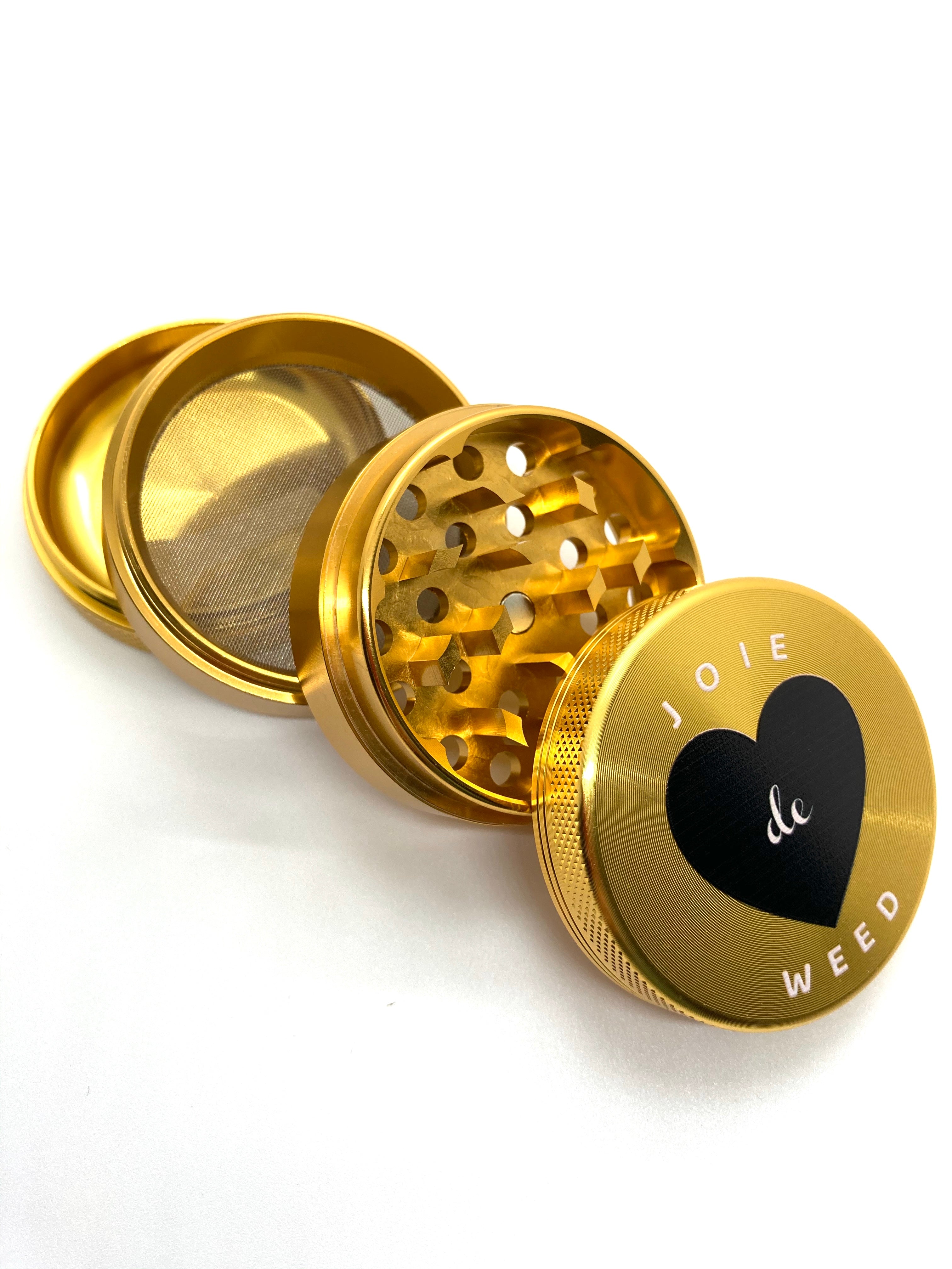 The 4-piece Joie de Weed grinder shown with its included triangular scraper tool. The set is presented as a professional-grade tool for the discerning smoker, designed to elevate the daily ritual with both function and style.