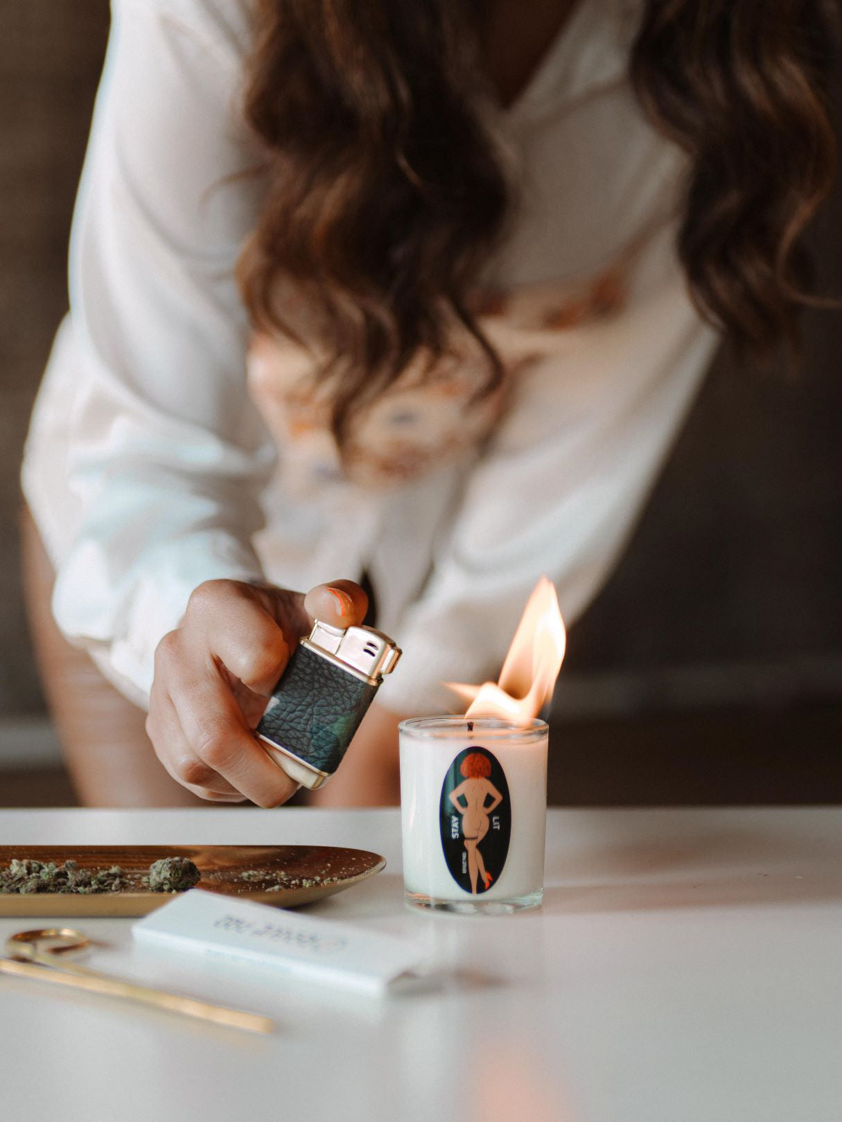 A shot of the 2oz clear glass votive candle being lit with a Rogue Paq refillable lighter. The smooth, off-white organic soy wax is visible, featuring a natural hemp wick. The label is clean and centered, emphasizing the high-design, apothecary-style aesthetic of the Rogue Paq aromatherapy collection. The label features the Rogue Paq signature can-can nude dancing girl with brown skin and red hair.