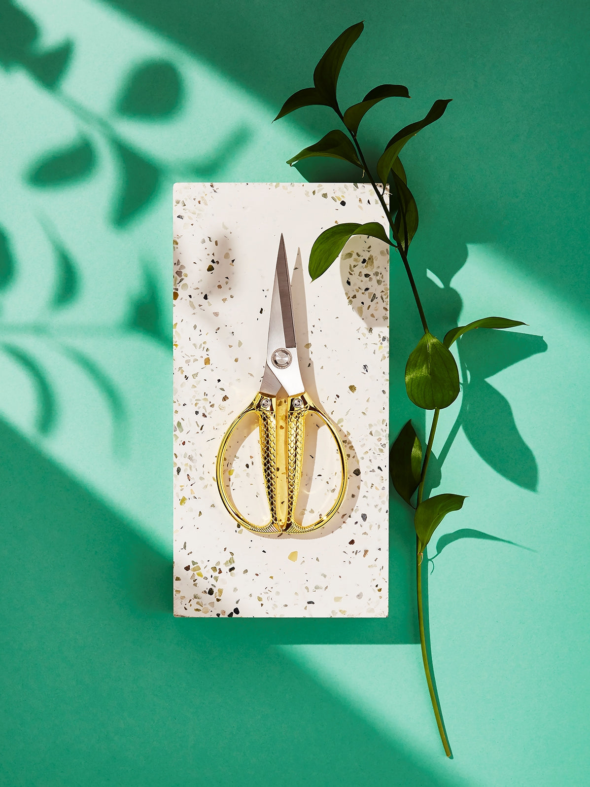 A pair of gold-toned stainless steel utility scissors with snake scale handles, displayed on a green background with a shadow falling over the scissors and some green leaves to the side.